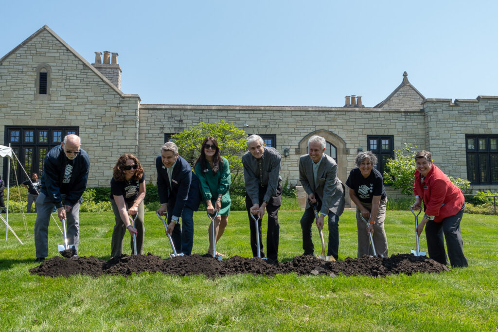 Eight people stand in a row with shovels in the dirt for the groundbreaking