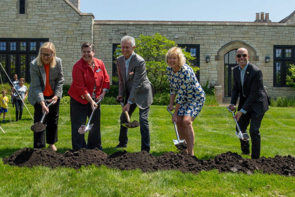 A group of five people stand in a row with shovels in the dirt for the groundbreaking.