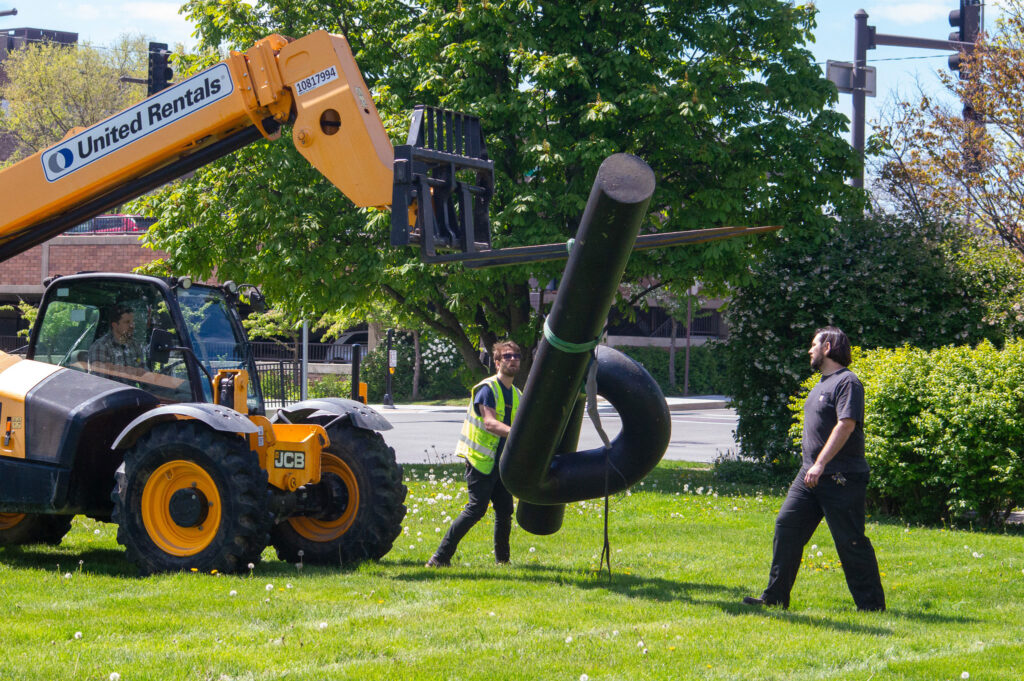 Two workers guide a piece of a sculpture that is suspended in the air by a tractor arm.
