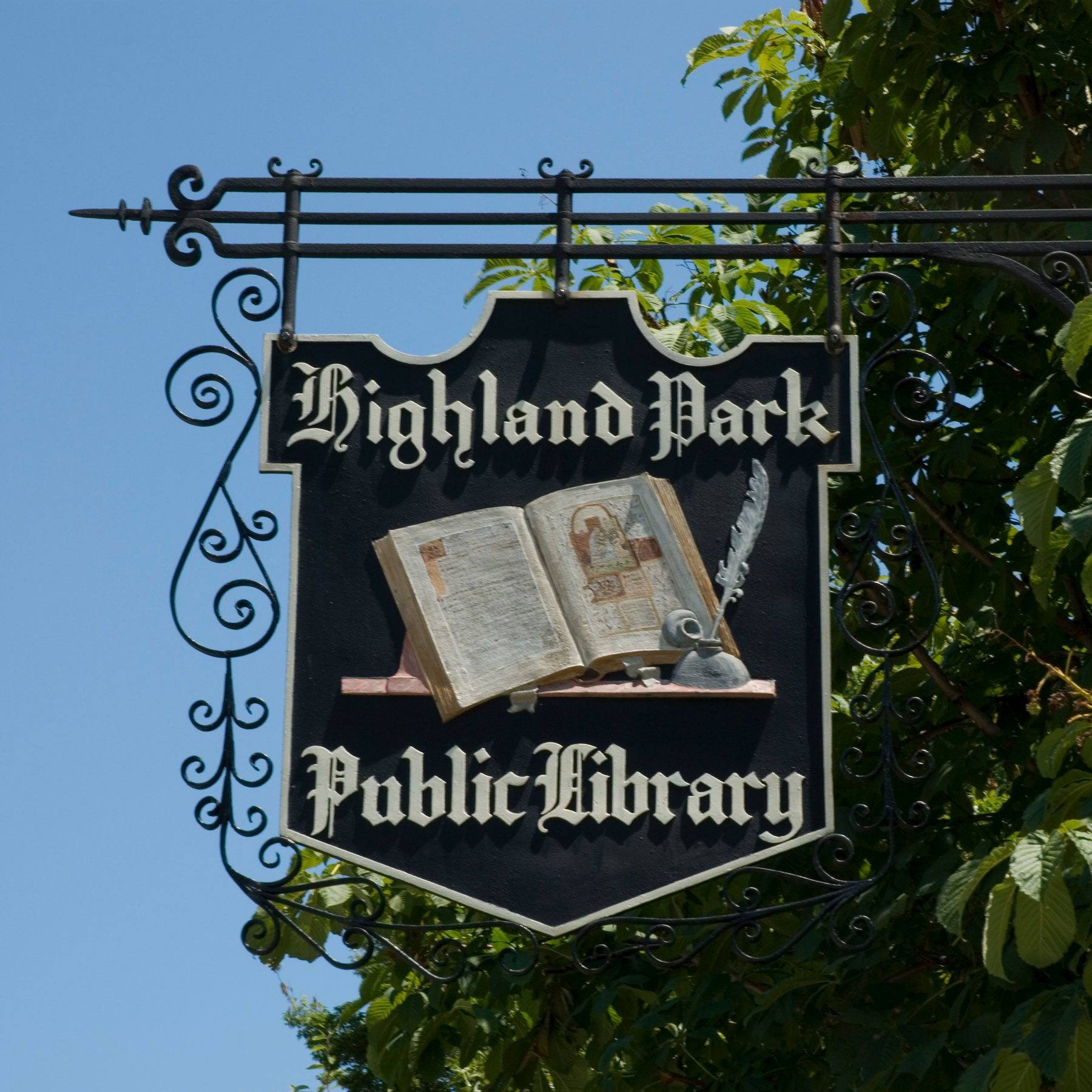 Detailed view of a hanging sign of wood and wrought iron scrolls. The name Highland Park Public Library and an image of an open book with quill pen are carved into the wood and painted.