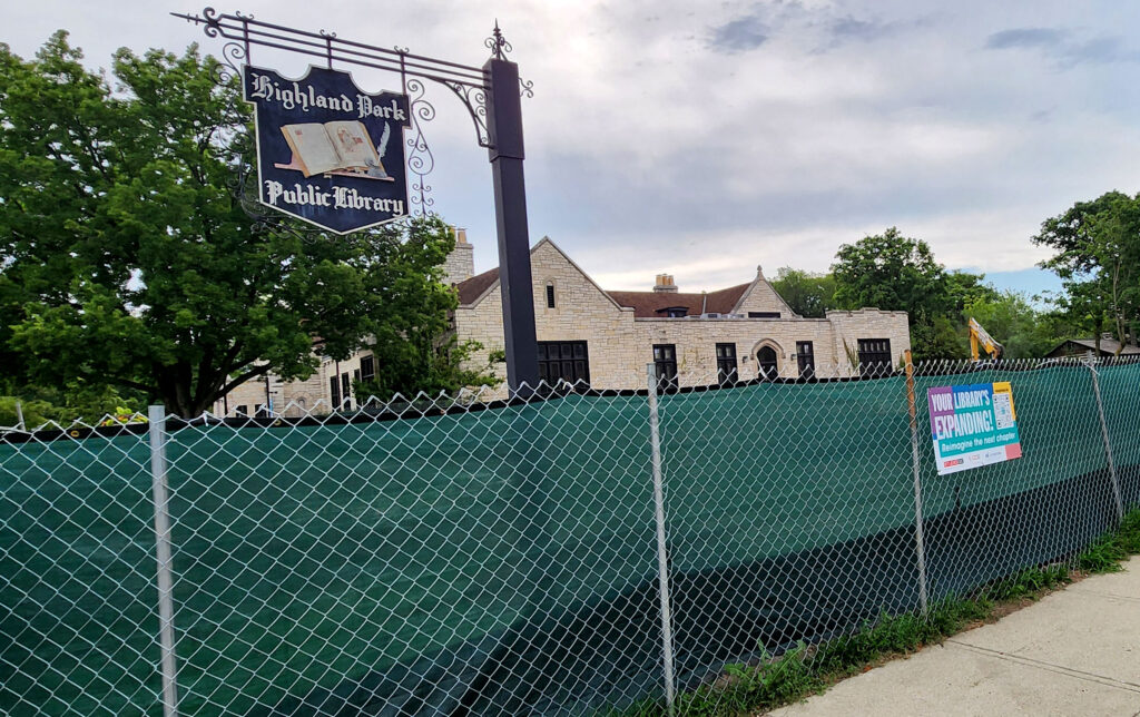 A hanging sign for Highland Park Public Library hangs from a pole from behind a construction fence. I sign on the fence says 