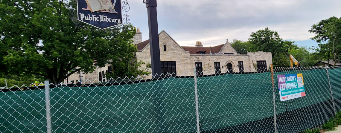 A hanging sign for Highland Park Public Library hangs from a pole from behind a construction fence. I sign on the fence says "Your Library's Expanding!" The west side of the library is visible behind the fence.