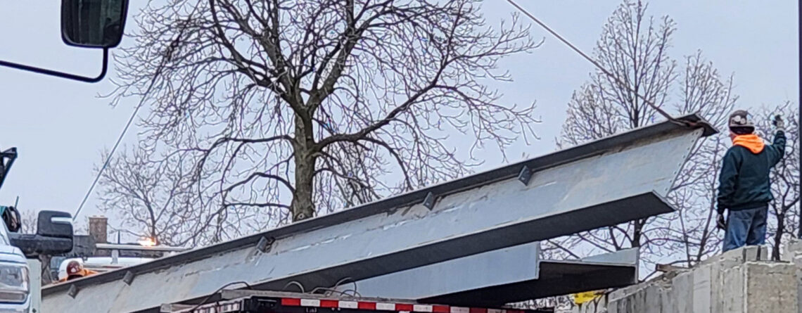 Steel girders are guided off of flatbed truck using a crane.