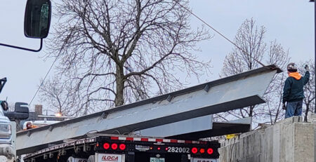 Steel girders are guided off of flatbed truck using a crane.